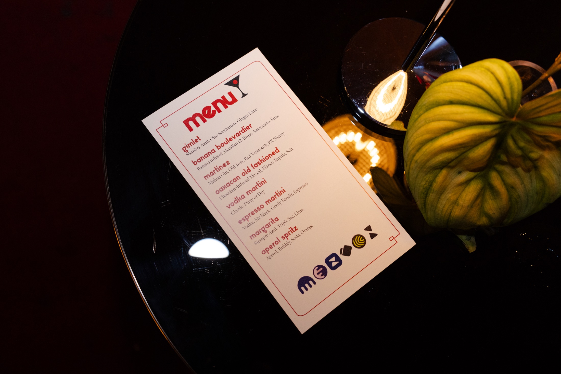 A printed cocktail menu with event partner logos beside a table lamp and green leaf