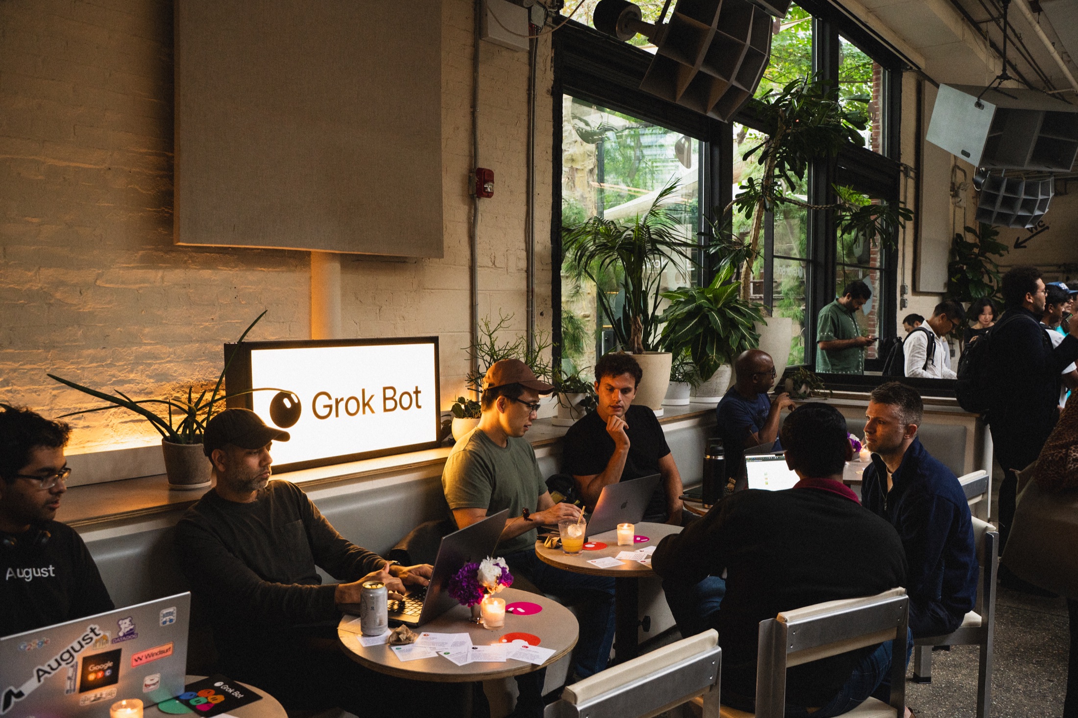Guests working on laptops and talking around tables beside an illuminated Grok Bot sign