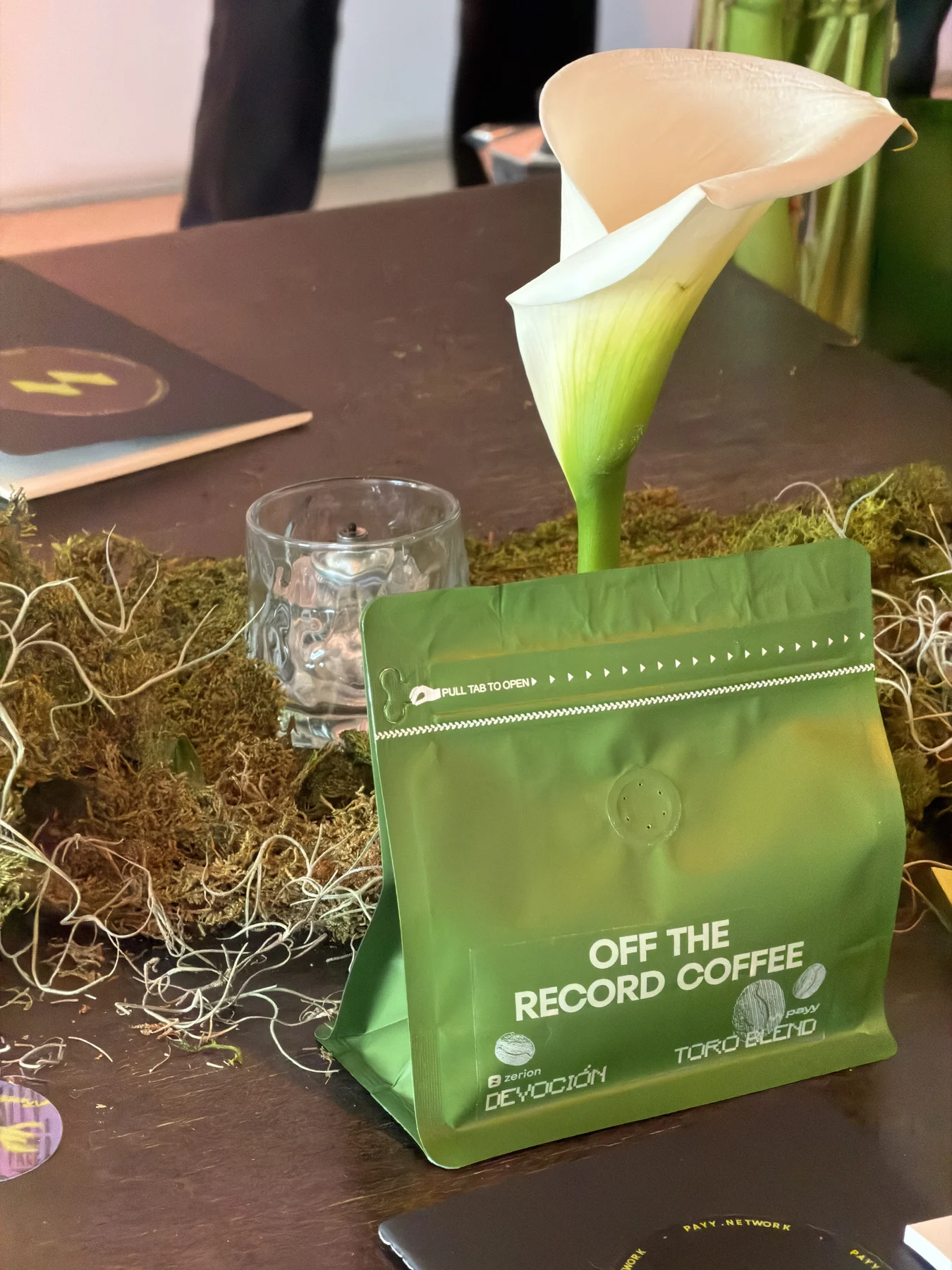 A green custom Off The Record Coffee bag from Devoción beside a calla lily and moss