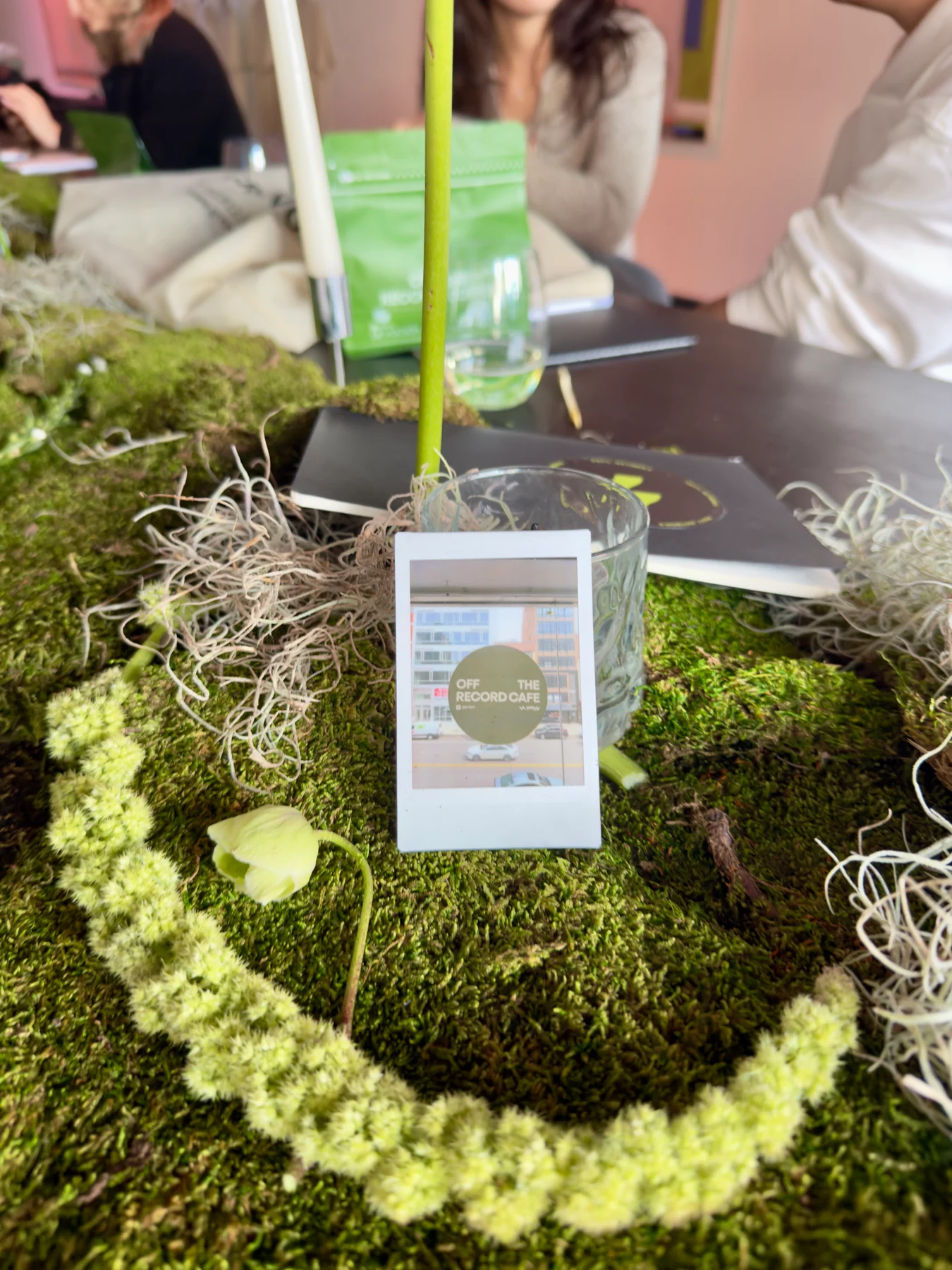 An instant photo of the Off The Record Cafe sign resting among moss, flowers, and branded notebooks
