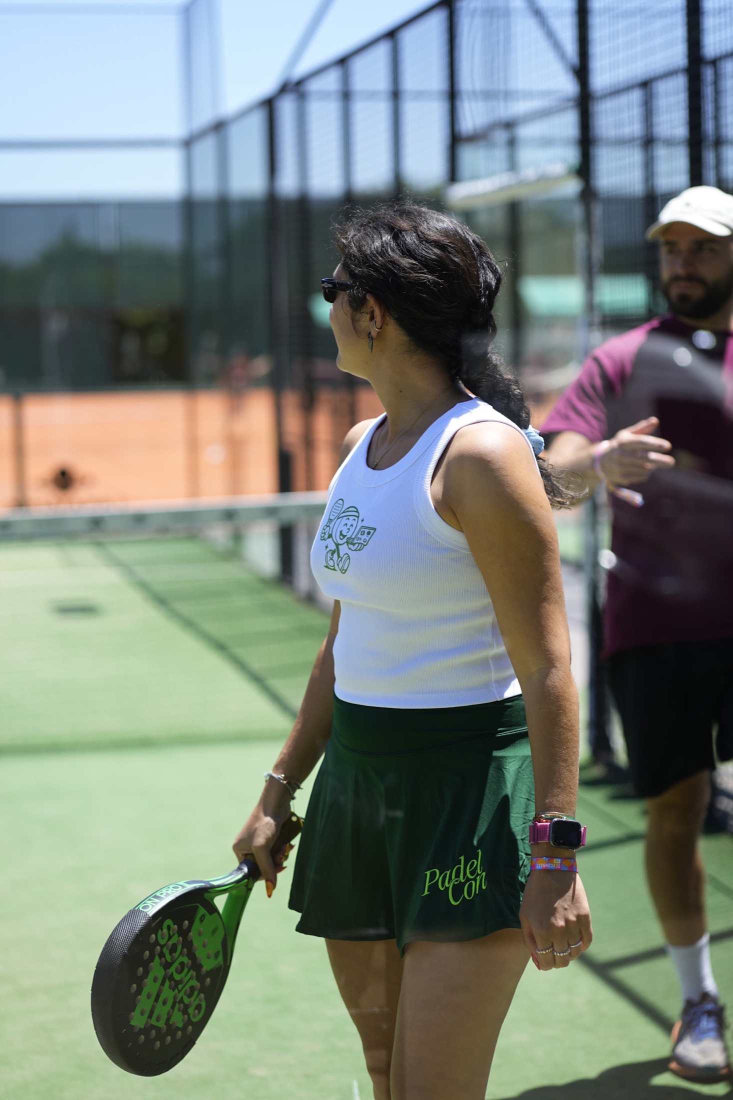 A player holding a padel racket and wearing a custom white tank top and green PadelCon skirt