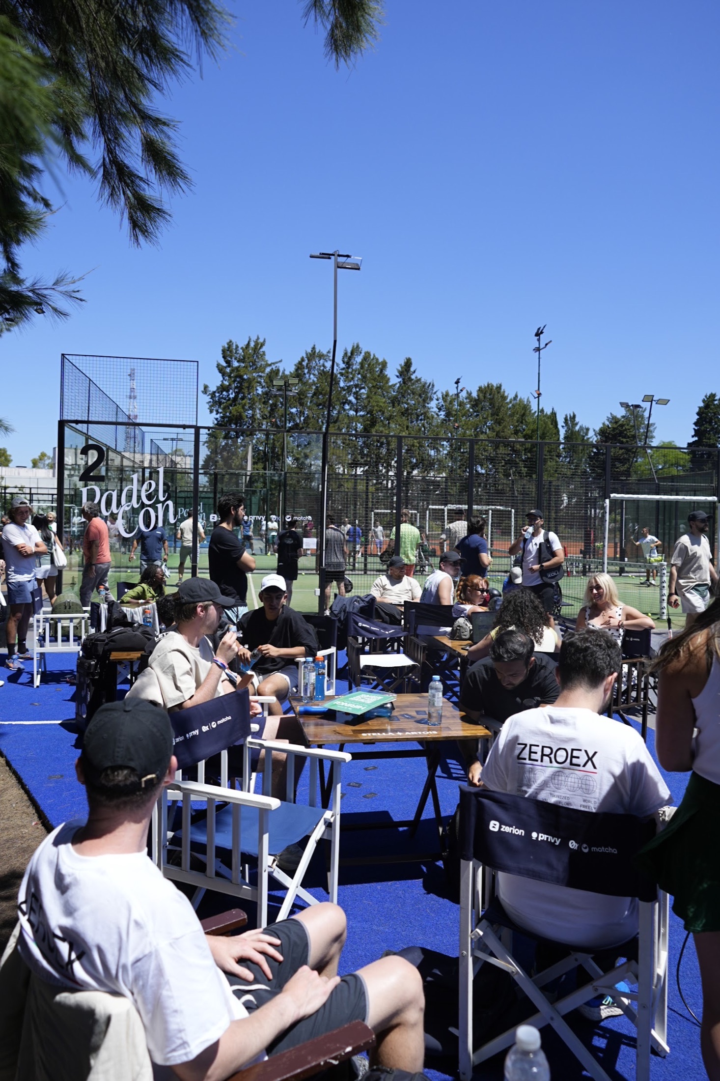 Guests seated around tables beside the PadelCon courts on a sunny day