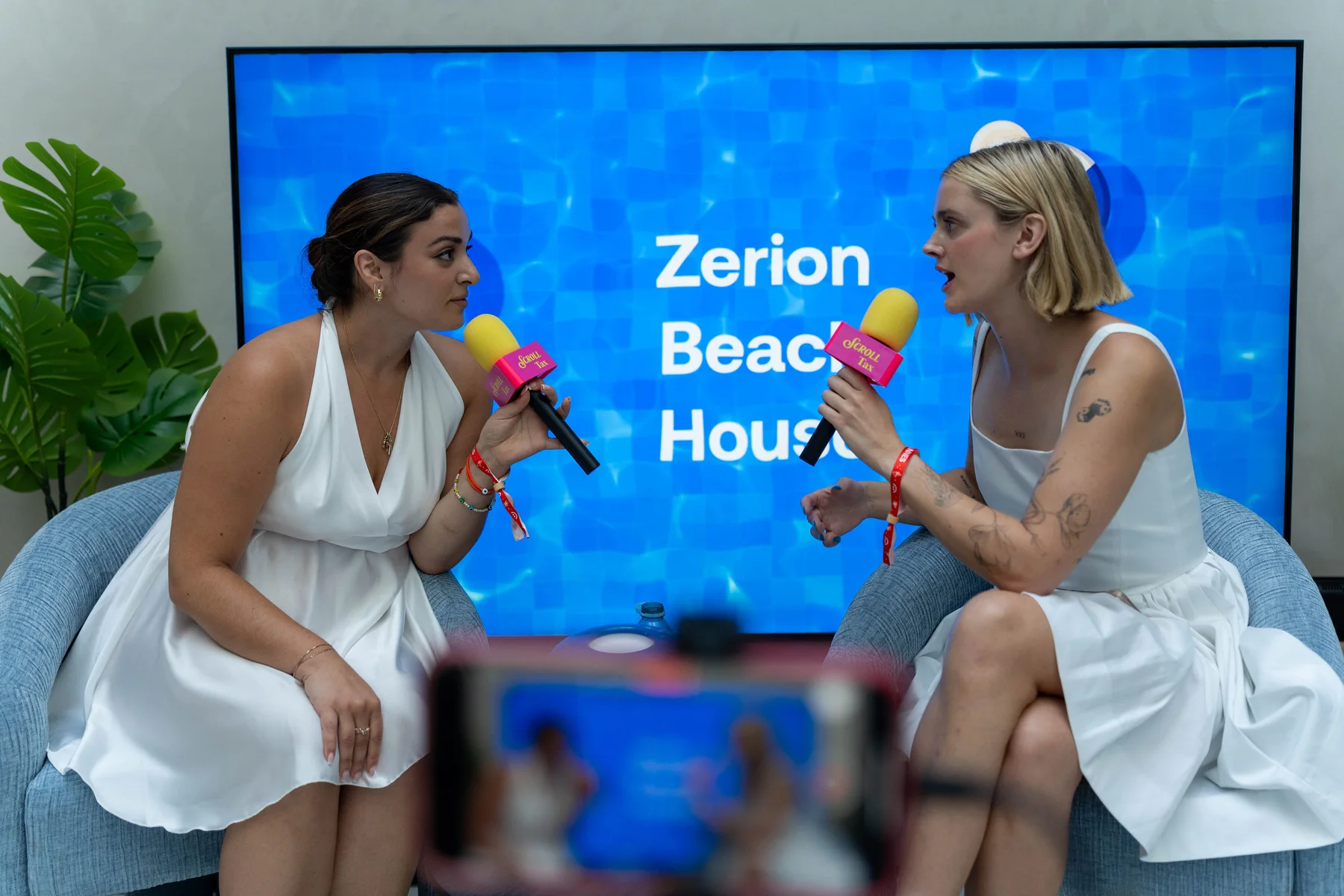 Two speakers with microphones seated in front of a blue Zerion Beach House screen, with a phone filming in the foreground